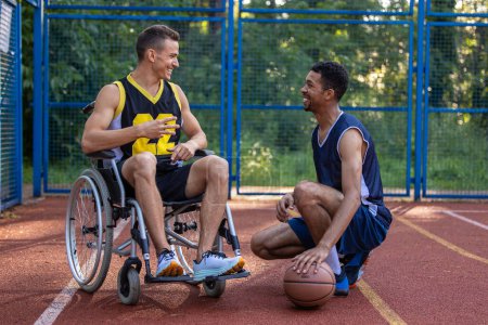 Photo for Caucasian man in wheelchair playing basketball with friend at outdoor court, talking while play game. - Royalty Free Image