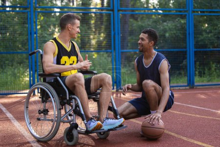 Foto de Caucásico hombre en silla de ruedas jugando baloncesto con un amigo en la cancha al aire libre, hablando mientras juega juego. - Imagen libre de derechos