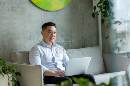 Téléchargez les photos : Asiatique homme gestionnaire de travail navigation sur le web dans son ordinateur portable tout en étant assis sur le canapé dans le bureau. - en image libre de droit