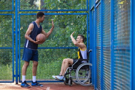 Foto de Caucásico hombre en silla de ruedas jugando baloncesto con un amigo en la cancha al aire libre, dando cinco. - Imagen libre de derechos