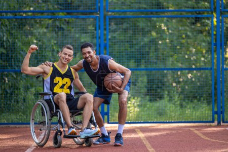 Foto de Caucásico hombre en silla de ruedas jugando baloncesto con un amigo en la cancha al aire libre, levantó el puño cerrado, mostrando poder. - Imagen libre de derechos