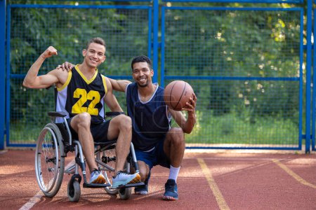 Téléchargez les photos : Homme jouant au basket avec un ami handicapé en fauteuil roulant au terrain extérieur. - en image libre de droit