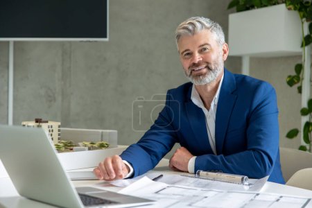 Photo for Grey haired man architect with beard working on some project, sitting at table in office in front of his laptop. - Royalty Free Image