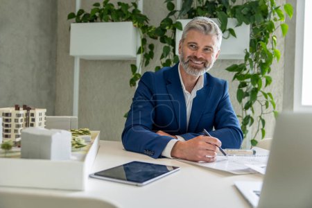 Téléchargez les photos : Homme aux cheveux gris architecte avec barbe travaillant sur un projet, assis à table au bureau devant son ordinateur portable. - en image libre de droit