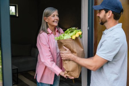 Téléchargez les photos : Courrier homme en bonnet livrant des légumes à la femme à la maison. - en image libre de droit