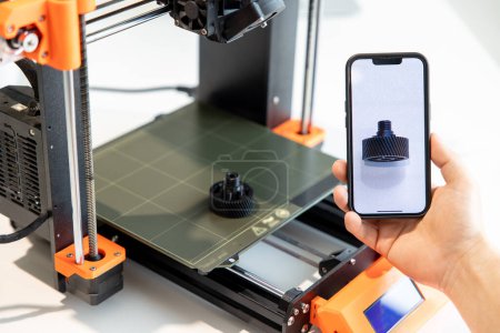 Téléchargez les photos : Homme méconnaissable tenant smartphone avec des détails à l'écran, travaillant sur une imprimante 3D dans un laboratoire créatif. - en image libre de droit