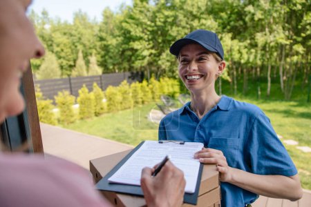 Photo for Smiling male courier delivering parcel, customer signs the delivery documents. - Royalty Free Image