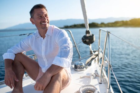 Foto de Feliz hombre. Hombre con ropa blanca sentado en una cubierta de yate y buscando relajado y feliz - Imagen libre de derechos