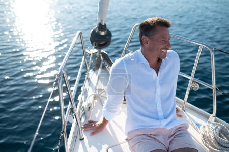 Téléchargez les photos : Un homme heureux. Homme en vêtements blancs assis sur un pont de yacht et regardant détendu et heureux - en image libre de droit