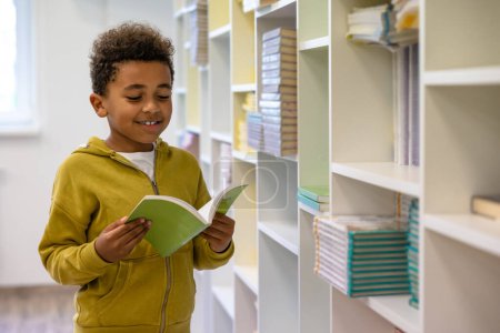 Foto de En la escuela. Lindo chico de piel oscura con capucha con un libro en las instalaciones de la escuela - Imagen libre de derechos