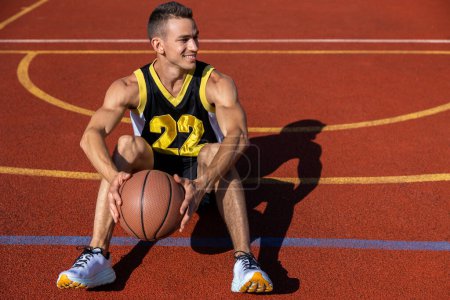 Photo for Smiling attractive man sitting on the basketball court after playing game, resting after match. - Royalty Free Image