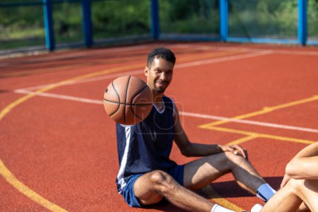 Foto de Alegre hombre atractivo descansando sentado en la cancha de baloncesto después de jugar, sosteniendo la pelota. - Imagen libre de derechos