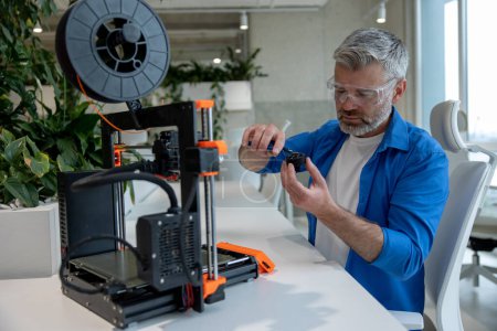 Téléchargez les photos : Équipement d'imprimante 3D moderne et homme mûr travaillant avec elle dans le bureau. - en image libre de droit