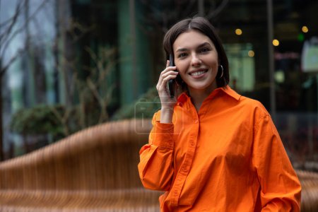 Foto de En la llamada. Chica de camisa naranja hablando por teléfono y sonriendo - Imagen libre de derechos