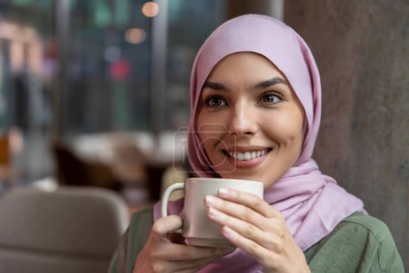 Téléchargez les photos : J'aime le café. Jolie jeune femme musulmane souriante appréciant son café - en image libre de droit