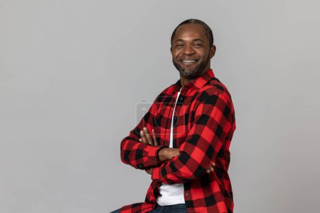 Téléchargez les photos : Homme barbu noir heureux confiant avec les mains pliées portant chemise à carreaux rouges posant isolé sur fond de studio gris. - en image libre de droit