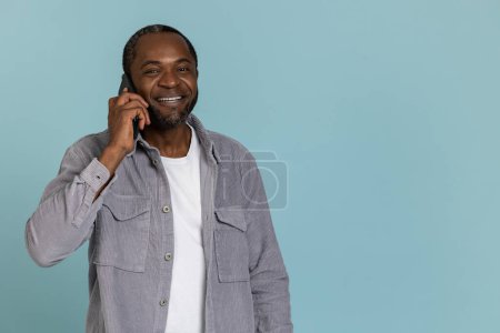 Foto de Feliz hombre negro con camisa gris hablando en el teléfono móvil de pie aislado sobre fondo azul - Imagen libre de derechos