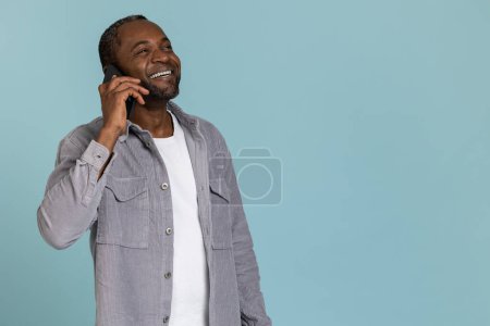 Foto de Encantado hombre negro con camisa gris hablando en móvil de pie aislado sobre fondo azul - Imagen libre de derechos