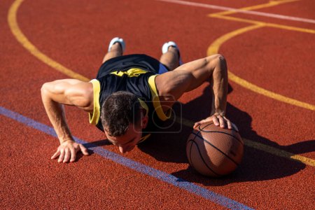 Téléchargez les photos : Beau jeune homme faisant des pompes avec une séance d'entraînement de balle de basket en plein air sur le terrain de basket. - en image libre de droit