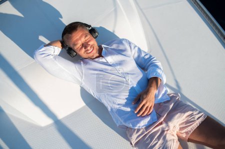 Foto de Feliz y relajado. Sonriente hombre con auriculares navegando en un yate y buscando relajado - Imagen libre de derechos