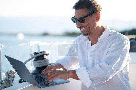 Téléchargez les photos : Sur internet. Homme souriant en lunettes de soleil passer du temps en ligne tout en naviguant sur un yacht - en image libre de droit