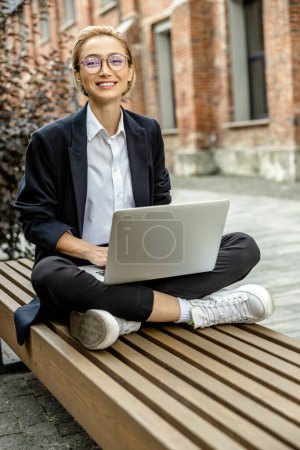 Téléchargez les photos : Je travaille. Jeune femme en lunettes assis avec les jambes croisées et travaillant sur un ordinateur portable - en image libre de droit