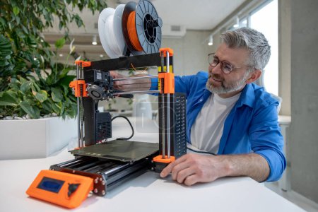 Téléchargez les photos : Équipement d'imprimante 3D moderne et homme mûr travaillant avec elle dans le bureau. - en image libre de droit