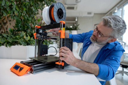 Foto de Equipo de impresora 3D moderno y hombre maduro que trabaja con él en la oficina. - Imagen libre de derechos