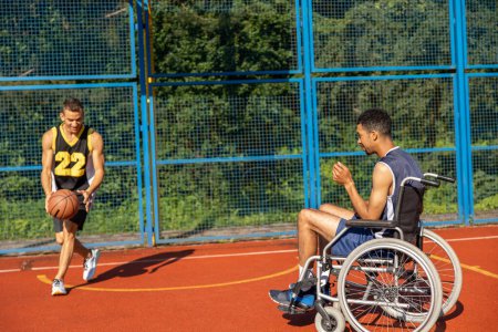 Téléchargez les photos : Homme handicapé en fauteuil roulant jouant au basketball avec un ami sur un terrain de sport. - en image libre de droit