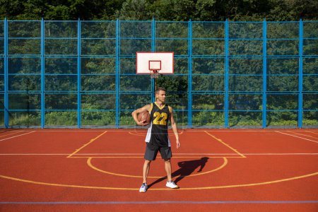 Téléchargez les photos : Jeune homme athlétique debout sur le terrain de basket-ball avec balle sous le bras. - en image libre de droit
