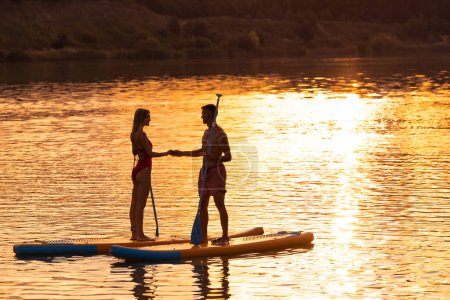 Téléchargez les photos : Silhouette de jeune femme romantique et homme surfant sur la planche SUP à travers la surface de l'eau. - en image libre de droit