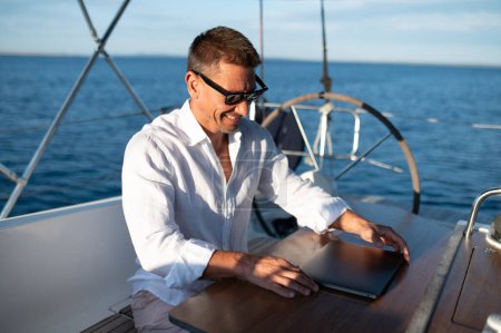 Téléchargez les photos : Nomade numérique. Homme confiant dans les lunettes de soleil travaillant sur un ordinateur portable tout en naviguant sur un yacht - en image libre de droit