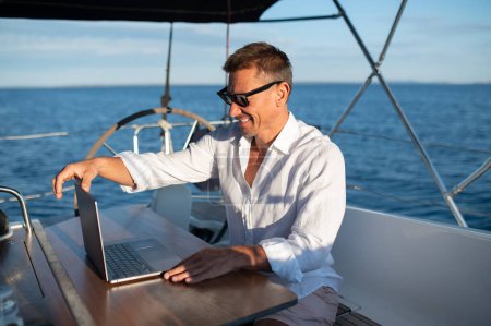 Digitaler Nomade. Selbstbewusster Mann mit Sonnenbrille arbeitet auf einer Jacht an einem Laptop