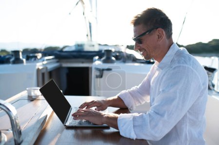 Digitaler Nomade. Selbstbewusster Mann mit Sonnenbrille arbeitet auf einer Jacht an einem Laptop