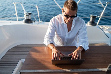 Photo for Work remotely. Man in white tshirt sitting on a yacht deck and typing on a laptop - Royalty Free Image