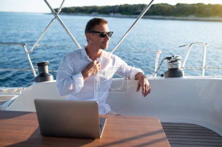 Photo for Work remotely. Man in white tshirt sitting on a yacht deck and typing on a laptop - Royalty Free Image