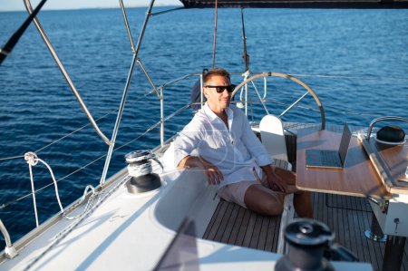 Téléchargez les photos : Café du matin. Satisfait homme en lunettes de soleil assis sur un pont de yacht avec une tasse de café - en image libre de droit