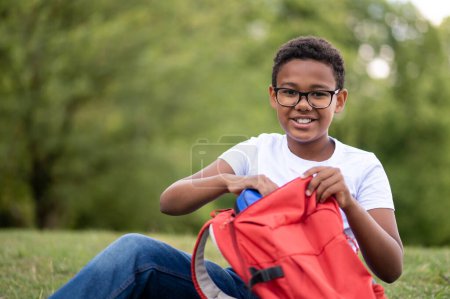 Téléchargez les photos : Souriant garçon. Un garçon avec un sac à dos rouge regardant heureux tout en étant assis sur le brut - en image libre de droit
