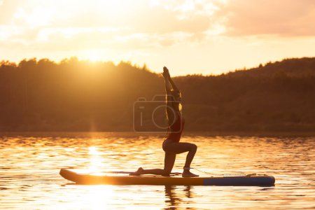 Foto de Mujer joven en postura de yoga practicando sobre tabla de sup paddle en mar tranquilo durante el amanecer o el atardecer. - Imagen libre de derechos