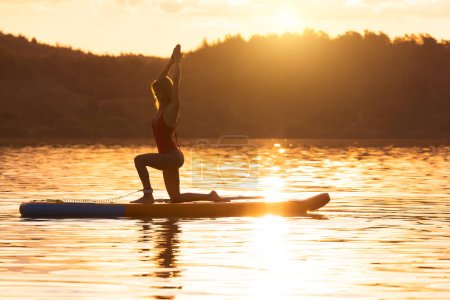 Téléchargez les photos : Belle femme pratiquant le yoga sur paddle sup surf au coucher du soleil. - en image libre de droit