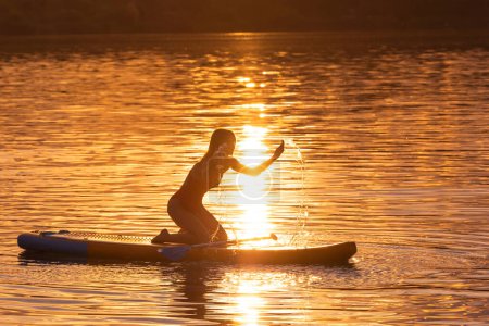 Téléchargez les photos : Silhouette de femme assise sur supboard au coucher du soleil profitant du surf. - en image libre de droit