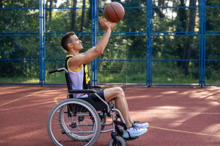Téléchargez les photos : Heureux joueur de basket-ball caucasien avec handicap utilise fauteuil roulant tout en jouant sur un terrain de sport extérieur. - en image libre de droit