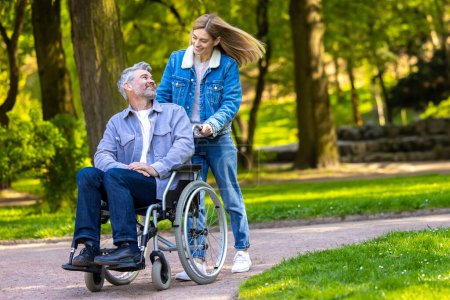 Téléchargez les photos : Bonne journée. Femme roulant un fauteuil roulant avec son mari dans le parc et les deux regardant heureux - en image libre de droit