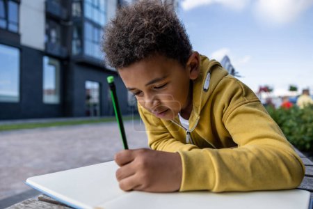 Foto de Haciendo los deberes. Colegial en sudadera con capucha amarilla mirando involucrado mientras hace la tarea - Imagen libre de derechos