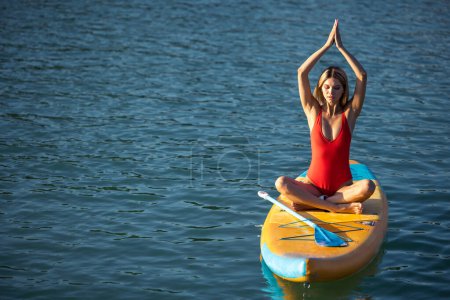 Téléchargez les photos : Blonde fille pratiquant le yoga pilates sur paddle sup planche de surf, femelle faisant de l'entraînement sur l'eau de mer. - en image libre de droit