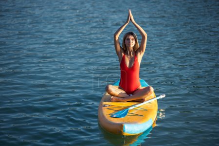 Téléchargez les photos : Jeune femme pratiquant le yoga sur paddle sup surf, activité sportive estivale individuelle féminine moderne. - en image libre de droit