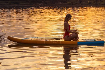 Foto de Mujer joven en postura de yoga practicando sobre tabla de sup paddle en mar tranquilo durante el amanecer o el atardecer. - Imagen libre de derechos