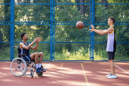 Téléchargez les photos : Athlète homme en fauteuil roulant et son ami s'amuser tout en jouant au basket-ball en plein air. - en image libre de droit