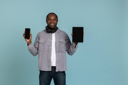 Téléchargez les photos : Attrayant jeune homme joyeux montrant tablette et smartphone écran vide isolé sur fond bleu. - en image libre de droit