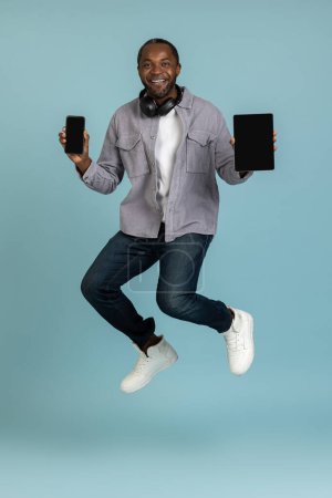 Foto de Atractivo joven alegre mostrando tableta y teléfono inteligente pantalla vacía saltando en el aire aislado sobre fondo azul. - Imagen libre de derechos